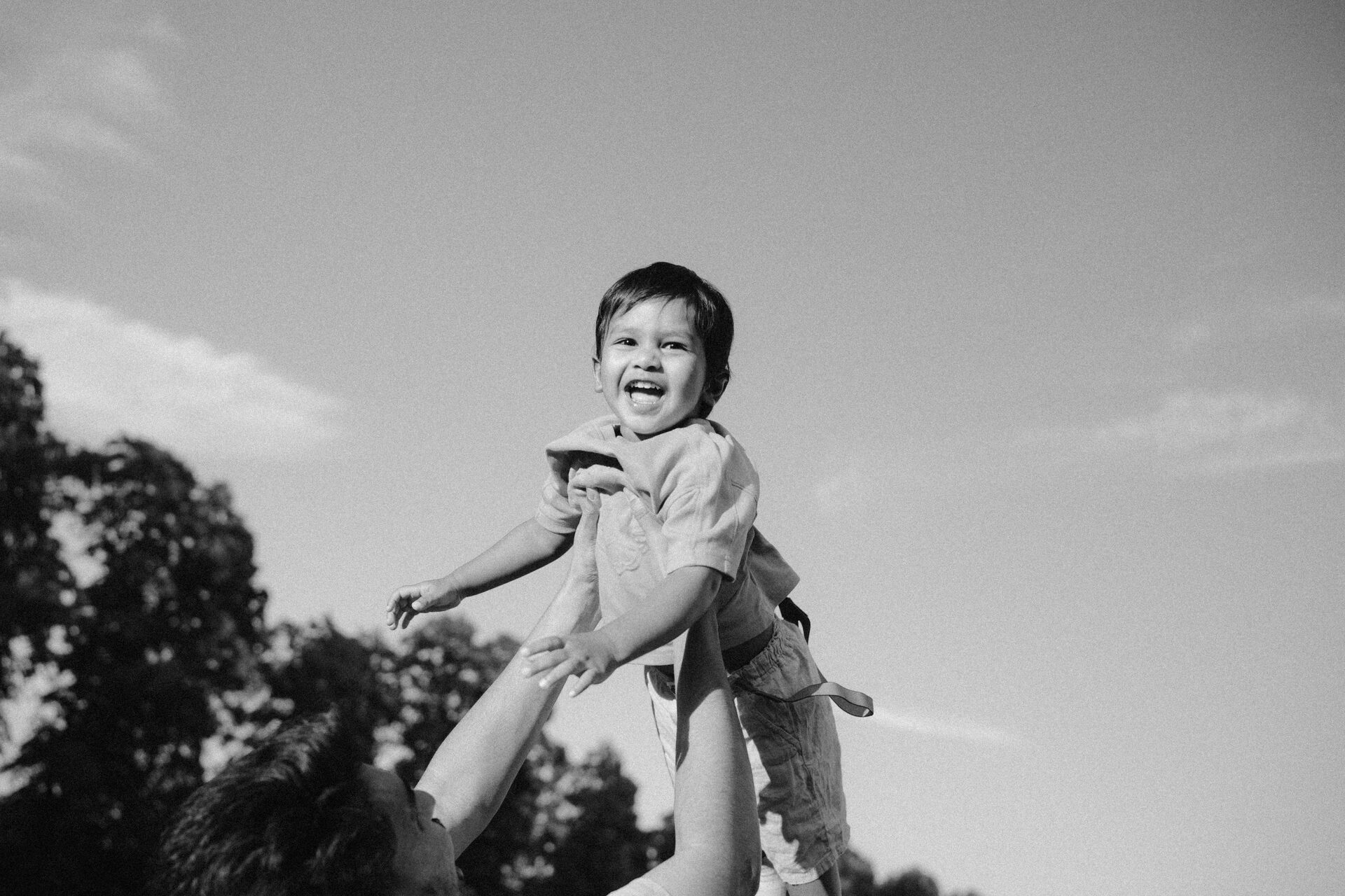 Black and white photograph of Aaru laughing while thrown into the air by his father at Victoria Park, Leicester — Mini Momentz