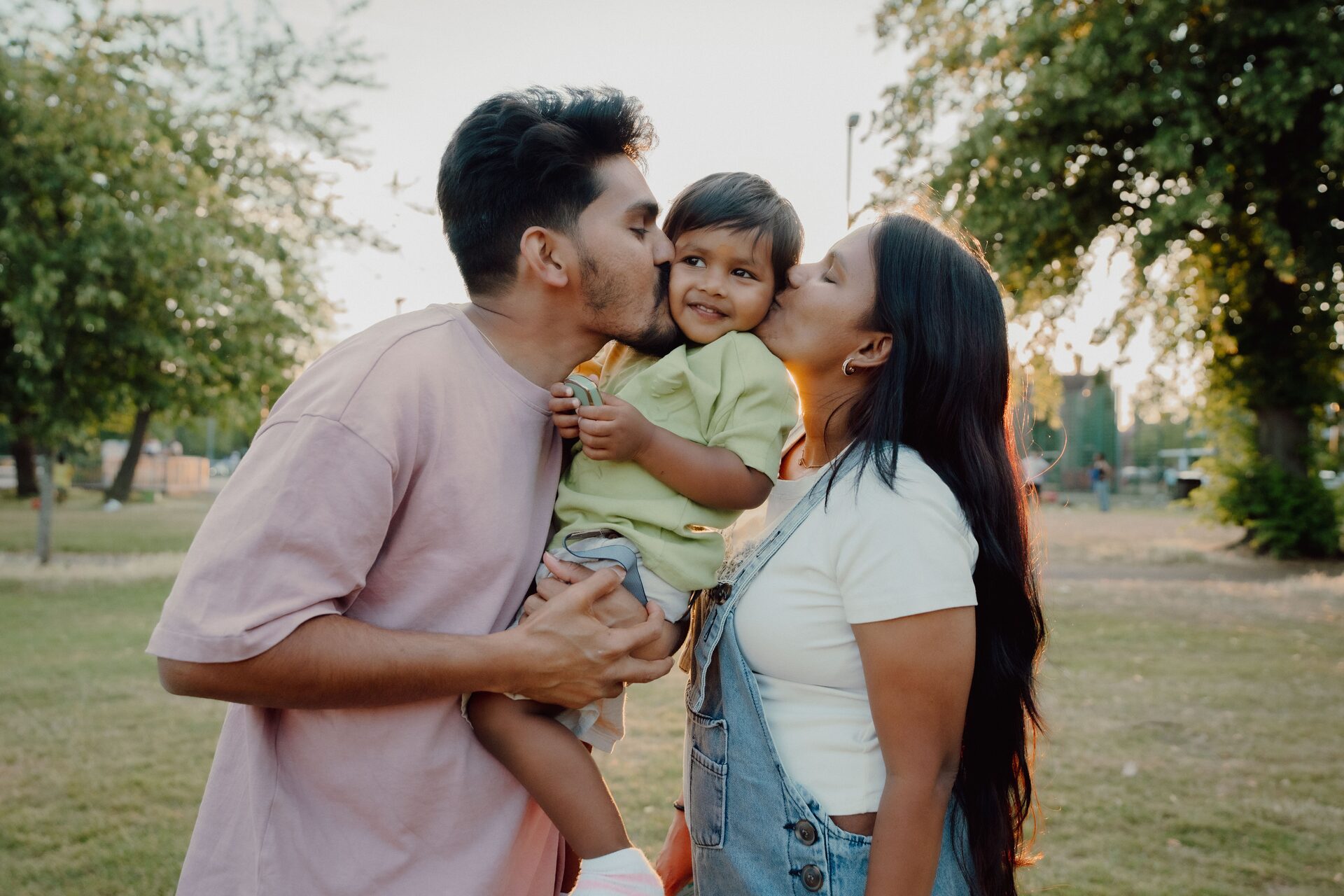 Harsha and Sravani kissing their son Aaru on the cheek at golden hour in Victoria Park, Leicester — Mini Momentz