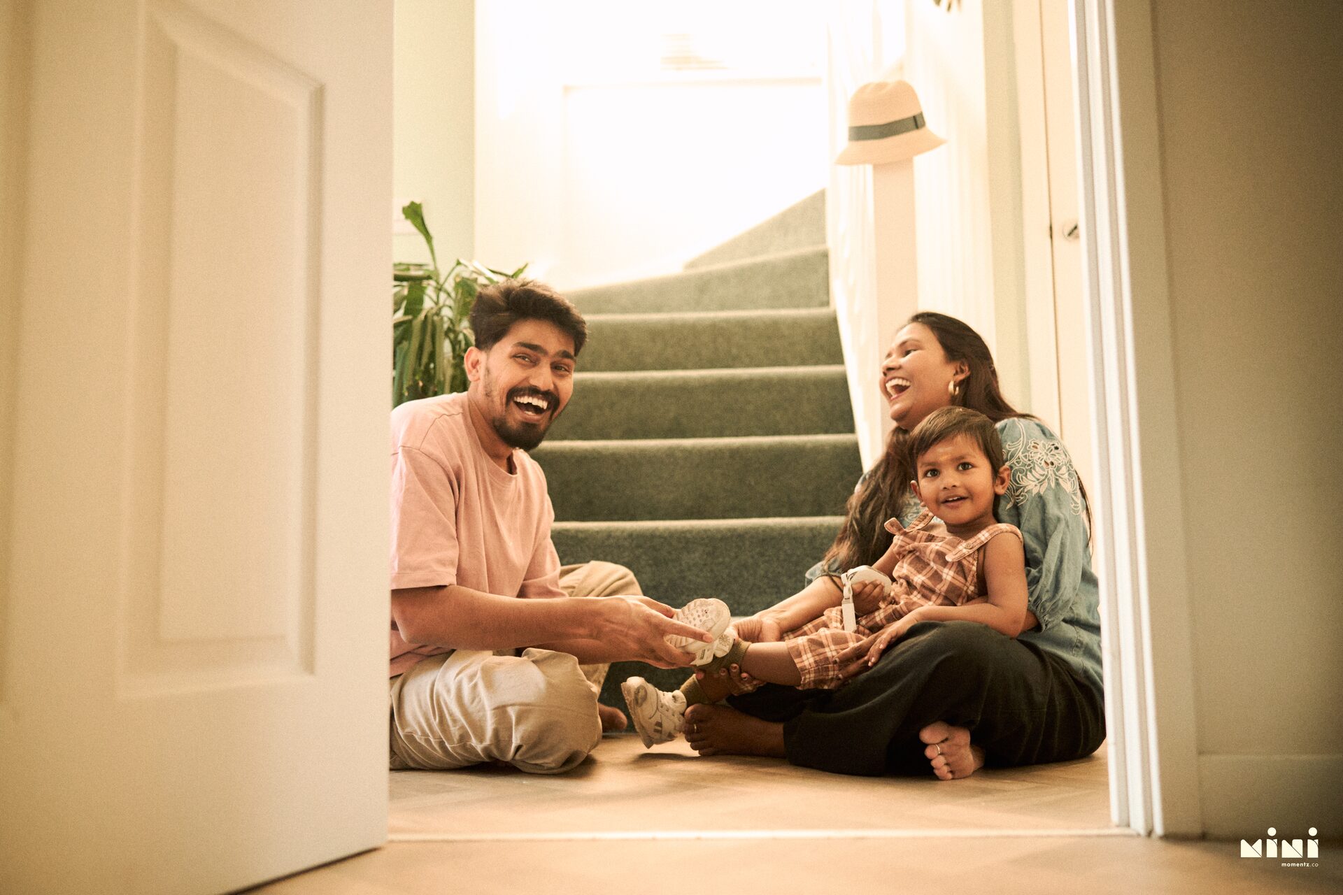 Harsha helping Aaru with his shoes at home in Leicester while Sravani laughs on the stairs — family photographer Leicester by Mini Momentz